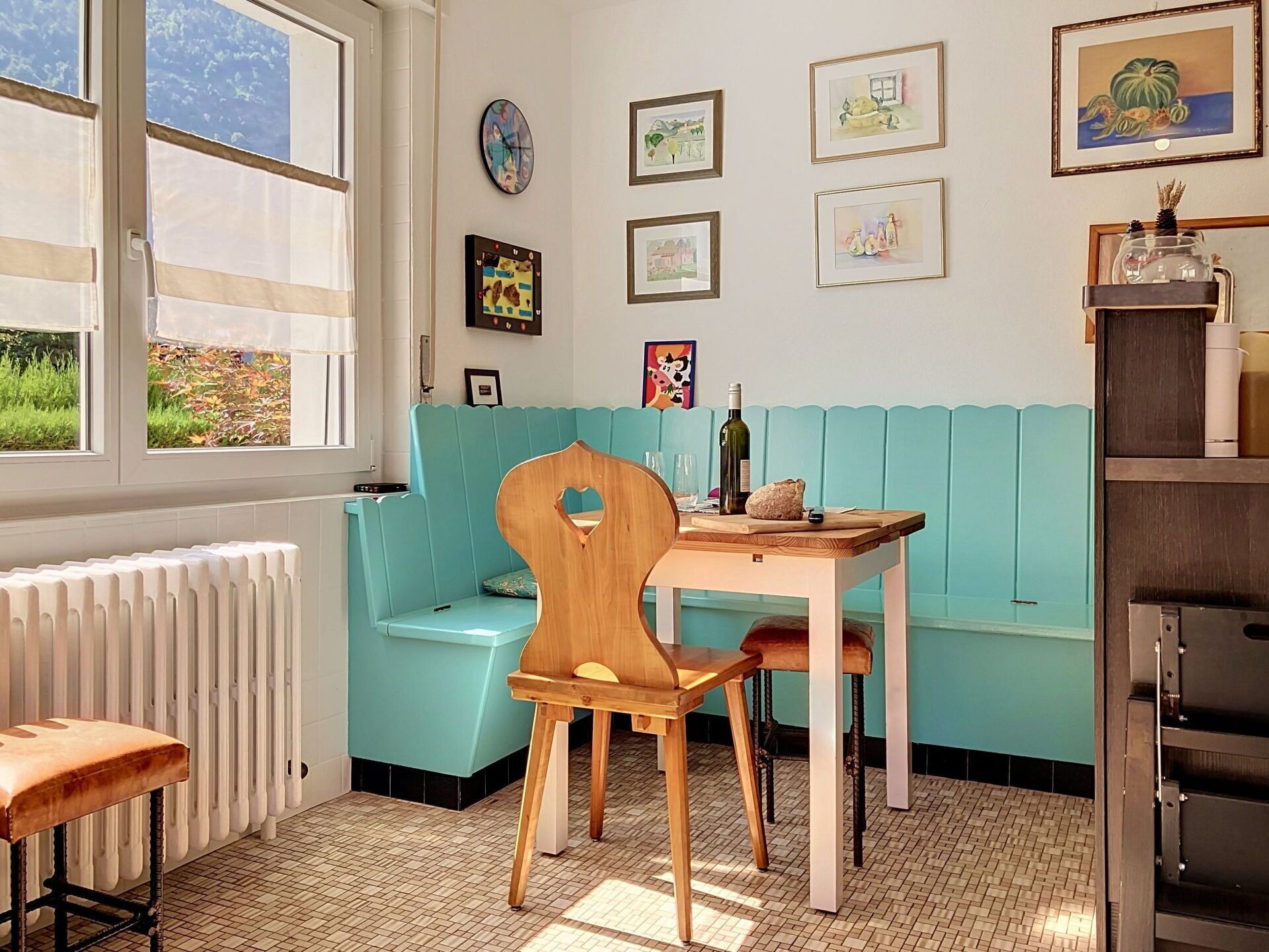Una stanza con una panca verde acqua, una sedia di legno, un tavolo con una bottiglia e una pagnotta di pane, e opere d'arte incorniciate appese al muro.