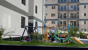 Um parquinho com balanços e estruturas de escalada, ao lado de um edifício residencial com vários andares.