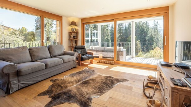 Ein modernes Wohnzimmer mit grauem Sofa, einem Holztisch, einem Ledersessel, einem Fellteppich und einem Balkon mit Blick auf Bäume.