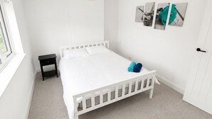 A white bedroom with a single bed, a small bedside table, a hanging lamp, and a window.