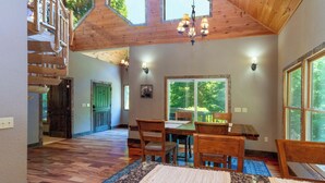 A spacious dining area with a high ceiling, wooden beams, and a large window offering a view of greenery. - Rookery-High-Rollers, Hiking & Hot Tubs–Ultimate Motorcycle-Friendly Retreat! (Whittier)