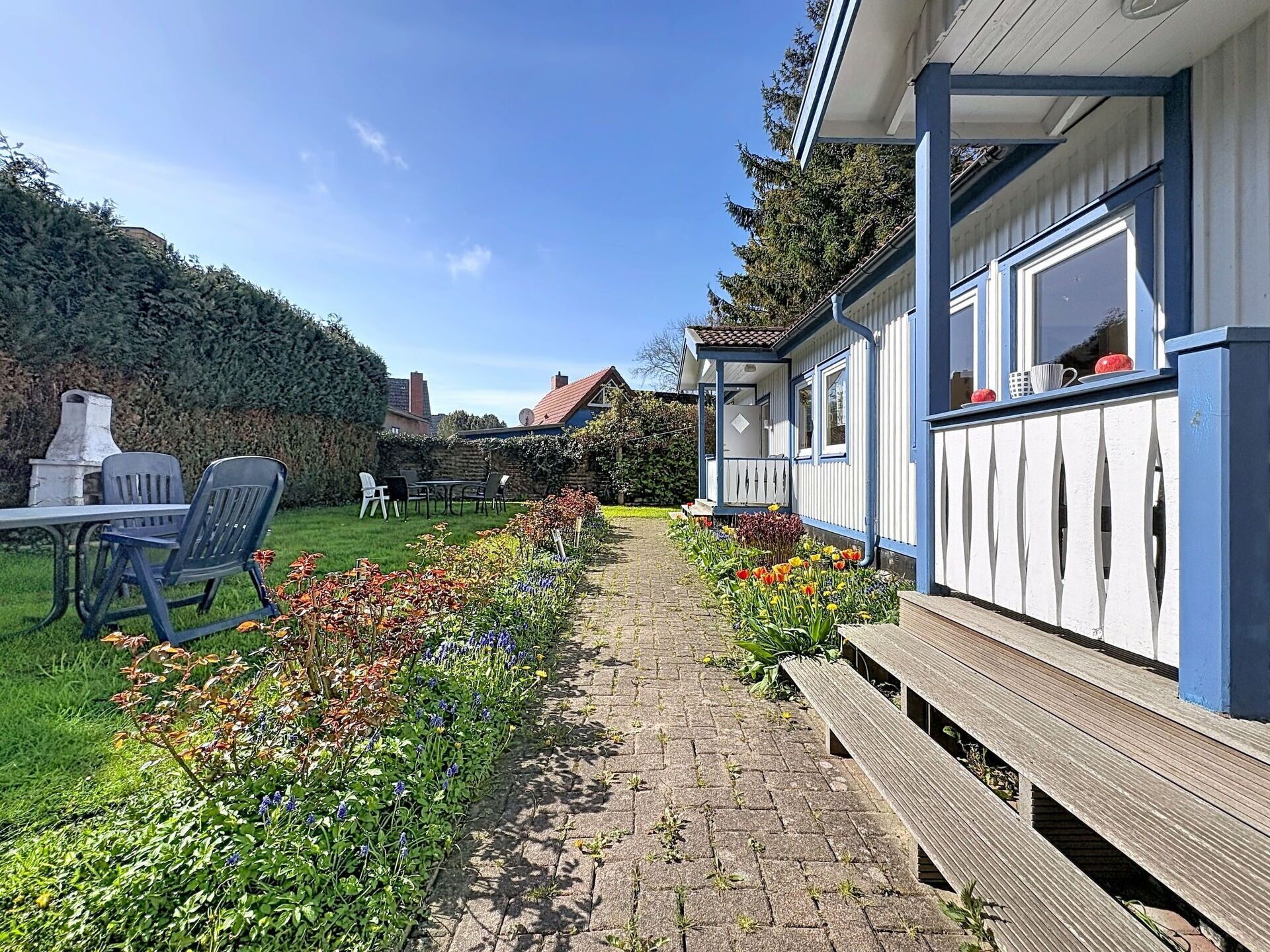 Ein Weg führt zu einem Gebäude mit einer Veranda, einem Garten mit Blumen und einem Tisch mit Stühlen.
