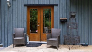 A wooden door with glass panels, two wicker chairs, a metal birdcage, and a small outdoor area with a wooden deck. - Rustic & Serene Forest 4 Bedroom Home - West Coast Retreat (Willis Point)