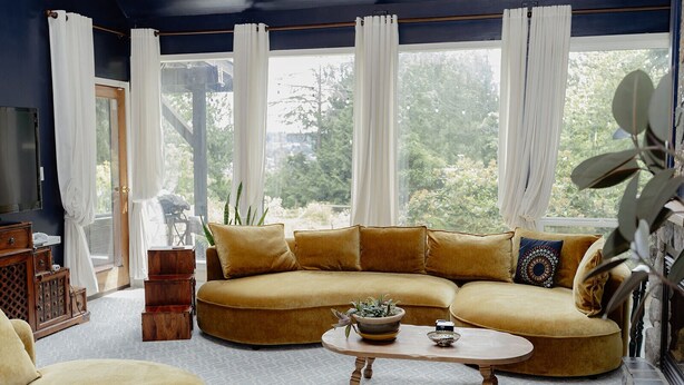 A living room with a curved sofa, a coffee table, and a TV. There are curtains, a plant, and a view of trees outside. - Rustic & Serene Forest 4 Bedroom Home - West Coast Retreat (Willis Point)
