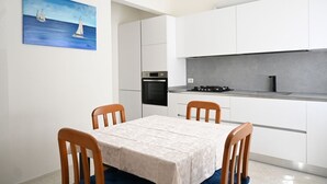 A modern kitchen with white cabinets, a stove, and a sink. There is a dining table with chairs and a painting of sailboats on the wall.