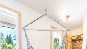 A room with a hanging hammock, a wooden floor, a grey sofa, and a view of trees through a window.