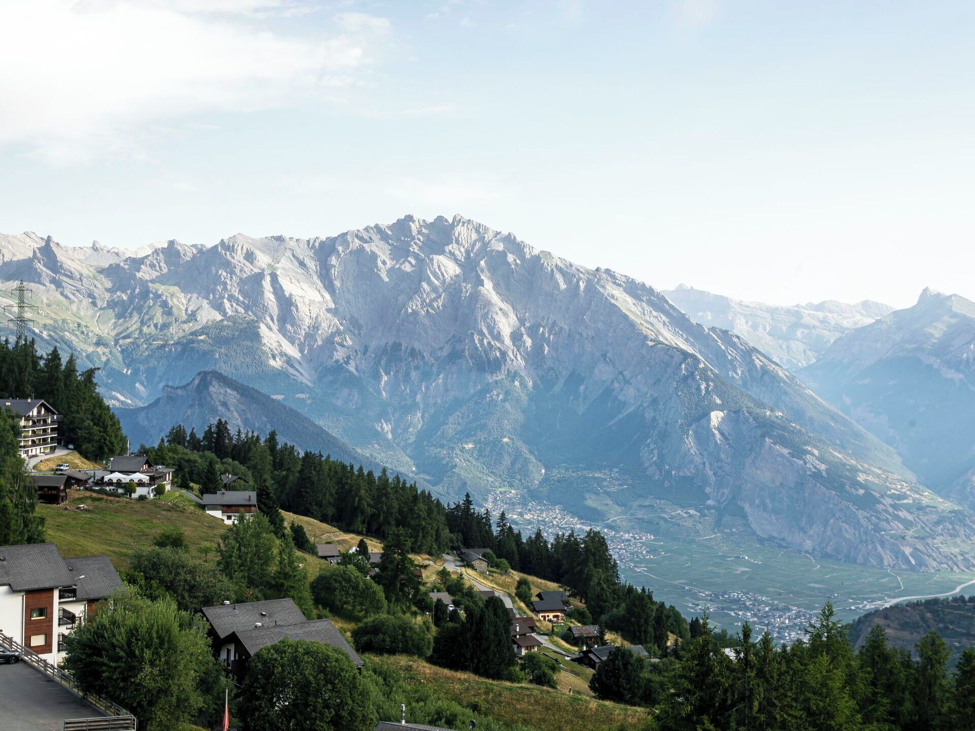 A mountain village with traditional houses, a valley, and snow-capped peaks in the background.