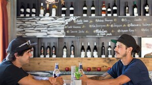 Twee personen zitten aan een ronde houten tafel met een kleine plant, in een bar met een muur vol wijnflessen en een krijtbordmenu.