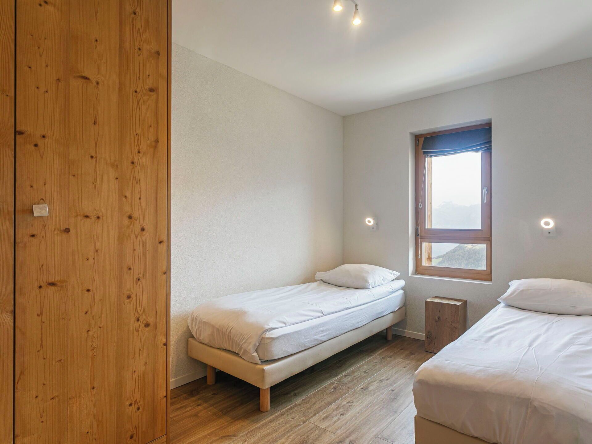 Une chambre avec une armoire en bois, un lit simple, un deuxième lit, une fenêtre donnant sur un beau paysage et une petite table de chevet.