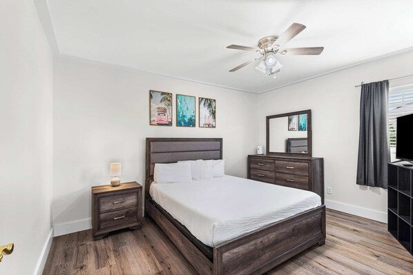 Un dormitorio con cama, mesitas de noche, ventilador de techo, obra de arte en la pared y una ventana con cortinas.