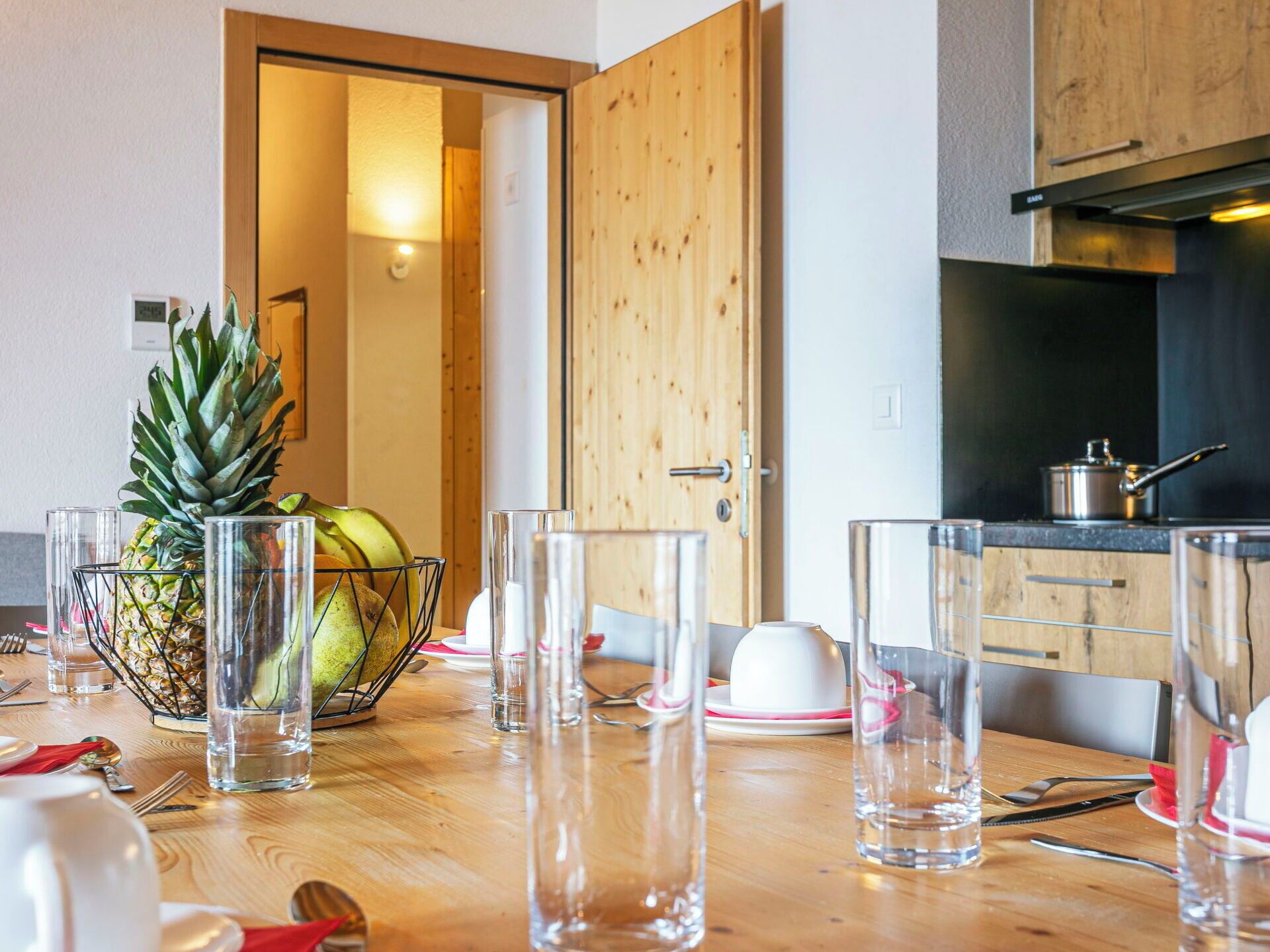 Une table à manger en bois, avec des verres transparents, un ananas et un panier de fruits.