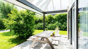 Patio esterno con tavolo e panche in legno, immerso nel verde e protetto da una struttura in vetro.