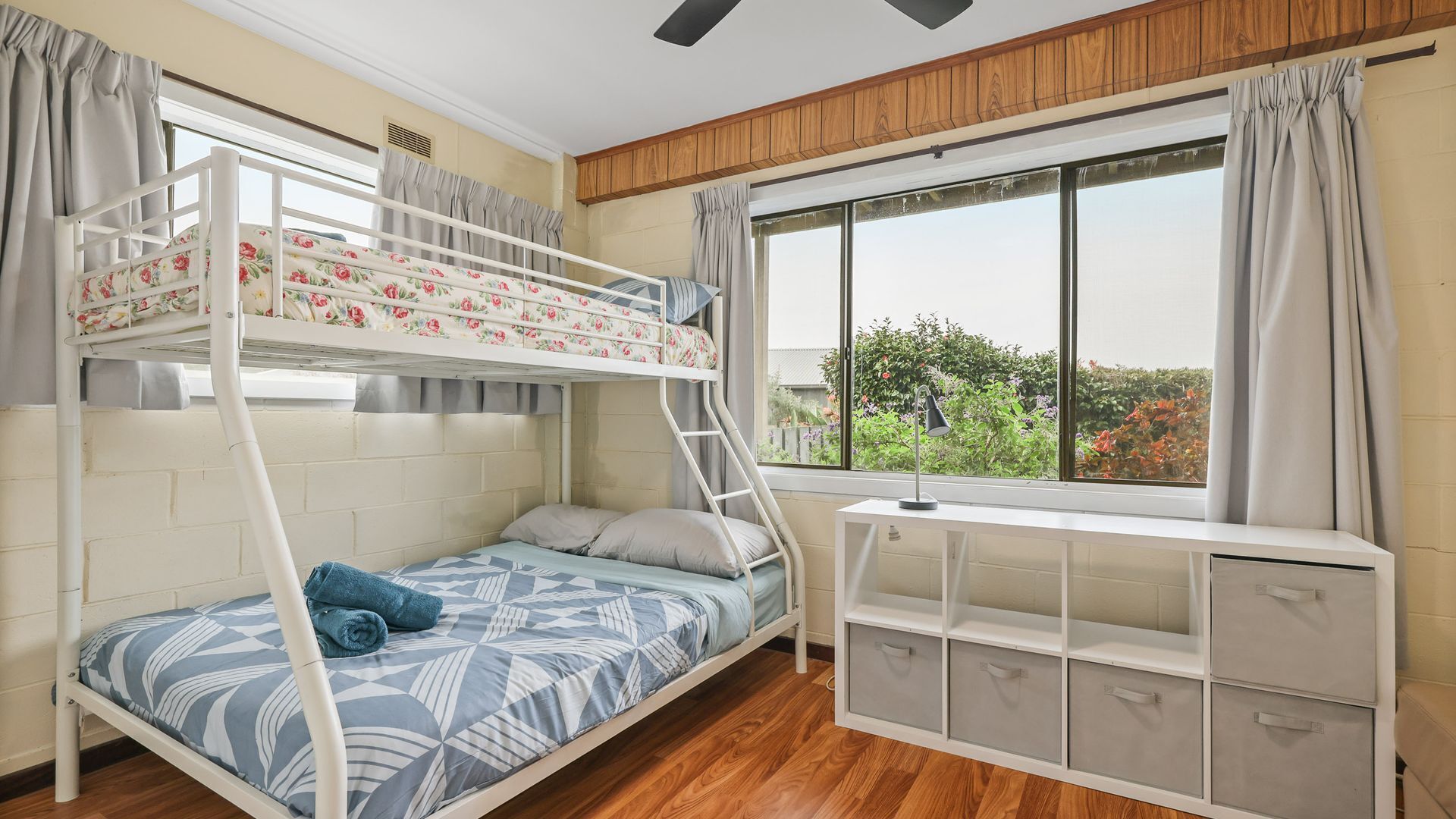 A bunk bed with floral bedding, a desk with storage drawers, a window with curtains, and a view of greenery outside.