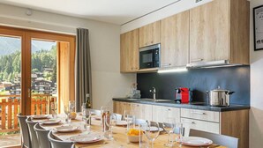 Een eetgedeelte met een houten tafel gedekt voor een maaltijd, een keuken met moderne apparatuur en uitzicht op de bergen door grote ramen.