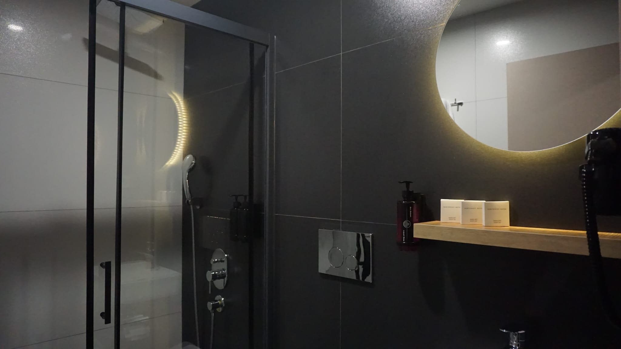 A modern bathroom with a glass shower enclosure, a round mirror, and a wooden shelf with toiletries.