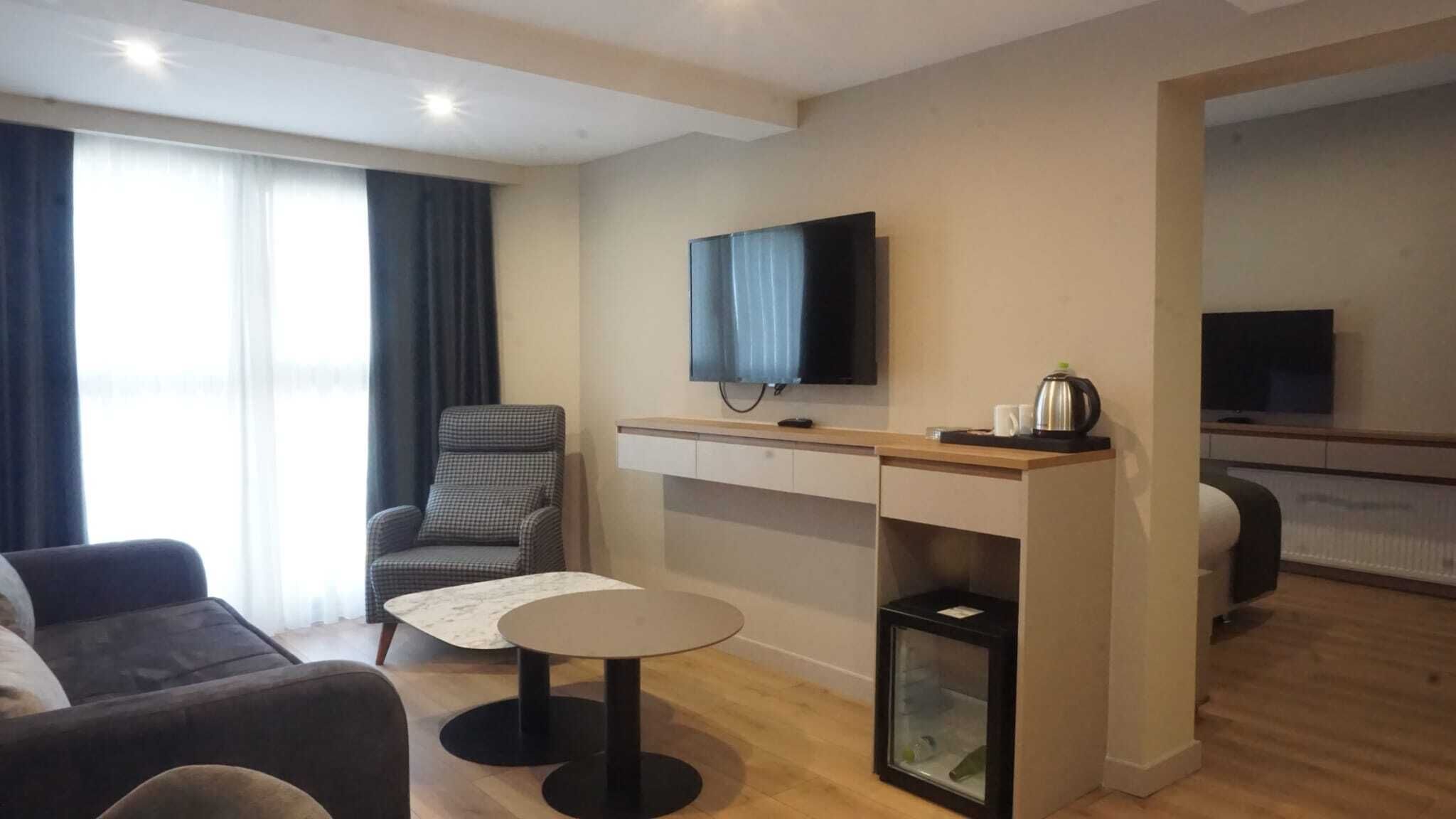 A modern hotel room with a sofa, a small round table, a TV mounted on the wall, and a kitchenette area.