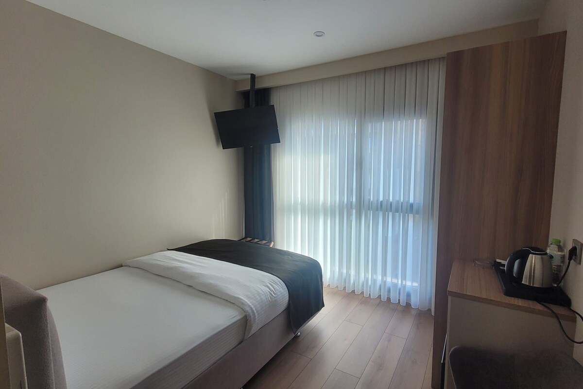 A hotel room with a bed, a TV mounted on the wall, a window with sheer curtains, and a small shelf with items.