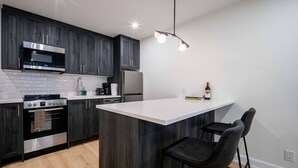 A modern kitchen with dark wood cabinets, stainless steel appliances, a white countertop island with bar stools, and a pendant light. - Modern 1BR in the Heart of Ottawa (Ottawa)