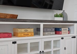 A shelf with board games, a television above, and a plant. - Heated Pool Access | Sleeps 10 | Steps to Beach (Gulf Shores)
