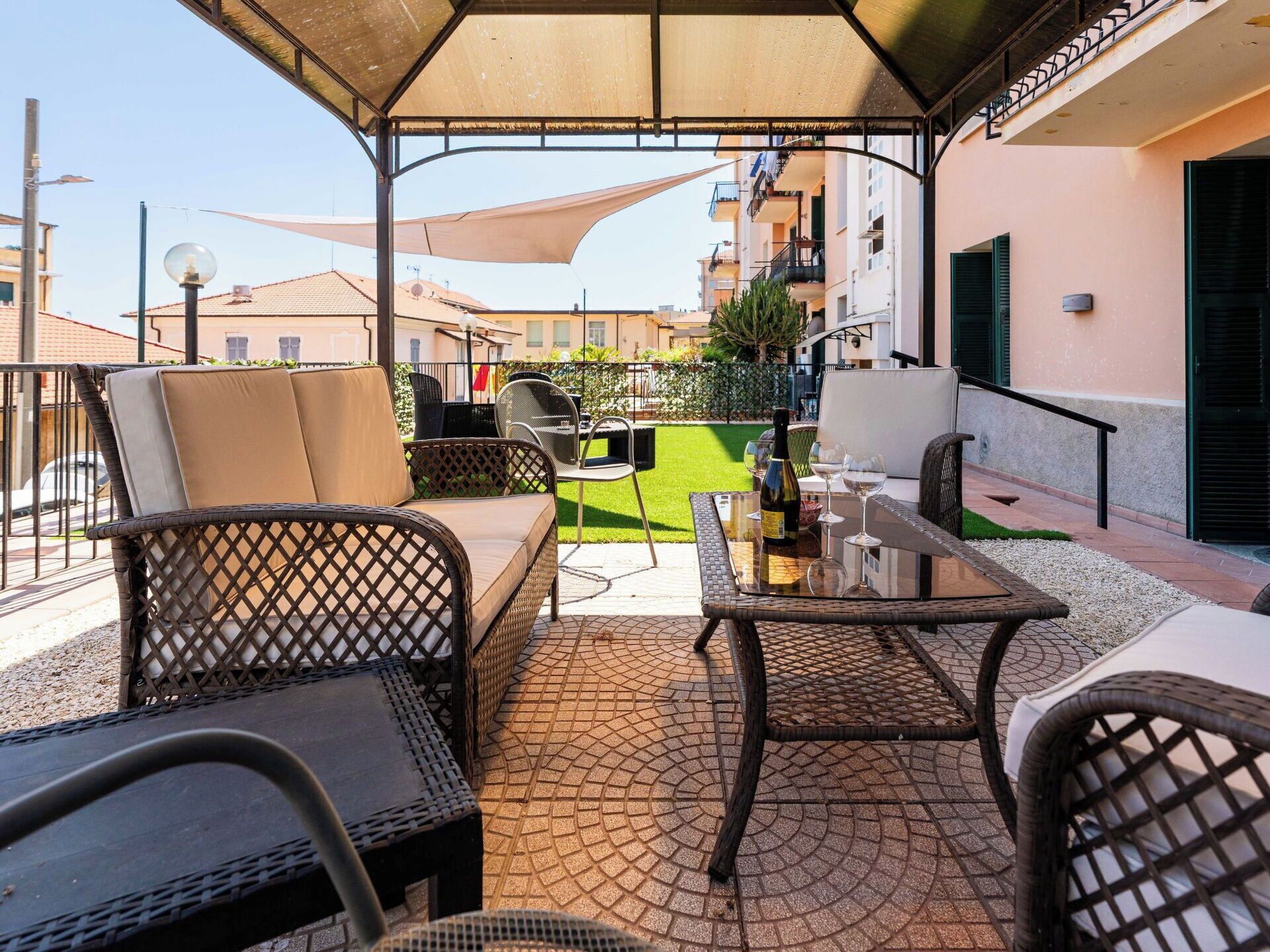 A covered outdoor seating area with wicker furniture, a table, and a bottle of wine.