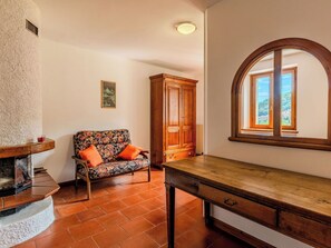 Una habitación con chimenea, una mesa de madera, un sofá con cojines naranjas, un aparador de madera y una ventana con vistas.