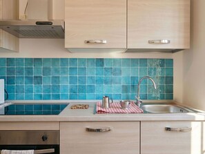 Eine Küche mit hellen Holzschränken, einer blauen Fliesenrückwand, einem Edelstahl-Spülbecken und einer kleinen Arbeitsplatte mit Wasserkocher und karierter Tischdecke.