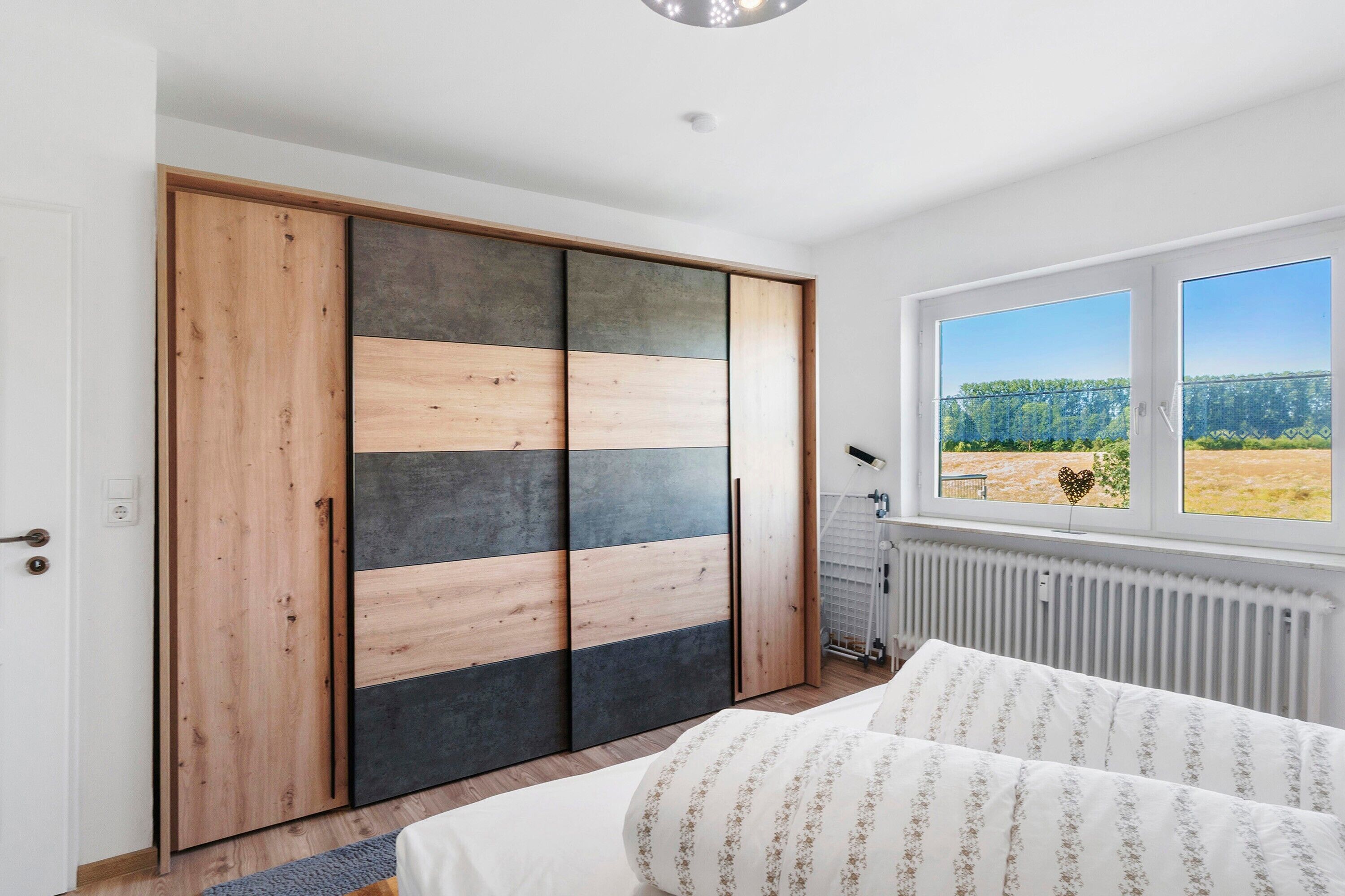 Un dormitorio con cama, armario de diseño en madera y color negro, radiador y ventana con vistas a zonas verdes.