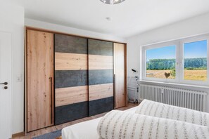 Une chambre à coucher avec un lit, une armoire dont le design combine des panneaux en bois et noirs, un radiateur et une fenêtre donnant sur un paysage verdoyant.