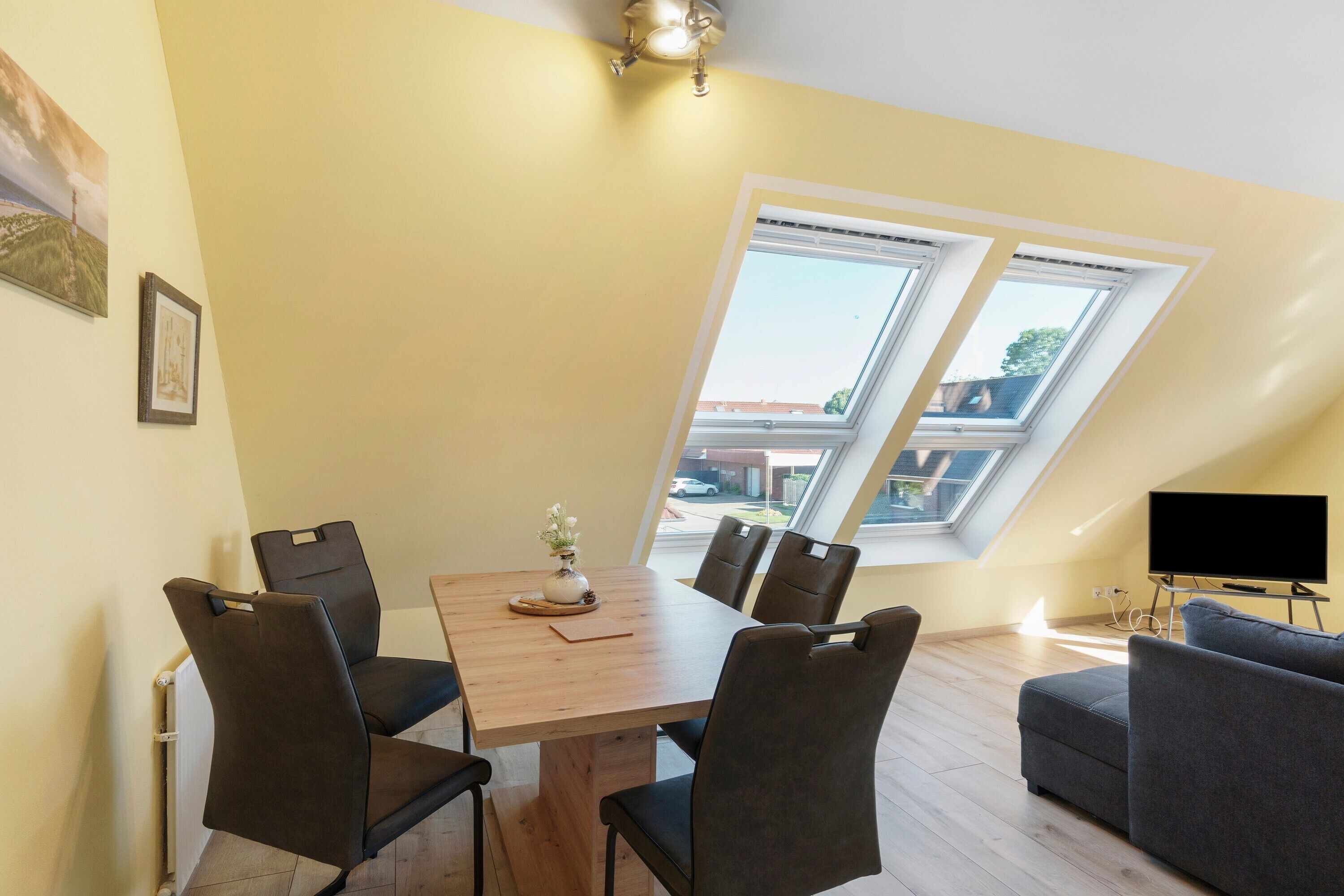 Ein Essbereich mit Tisch, Stühlen und einem Blumenstrauß. Zwei Fenster bieten einen Blick nach draußen.