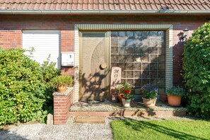 Une maison en briques avec une grande fenêtre, une porte surmontée d’un tympan et des plantes en pot.