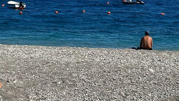 Une plage de galets bordée d’une mer bleue et limpide, avec des bateaux et une personne assise sur le rivage.