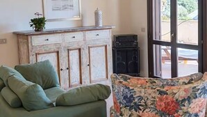 Un salon avec un canapé vert, un fauteuil à motifs floraux, une commode ornée d’un tableau encadré et une vue sur l’extérieur à travers une porte vitrée.