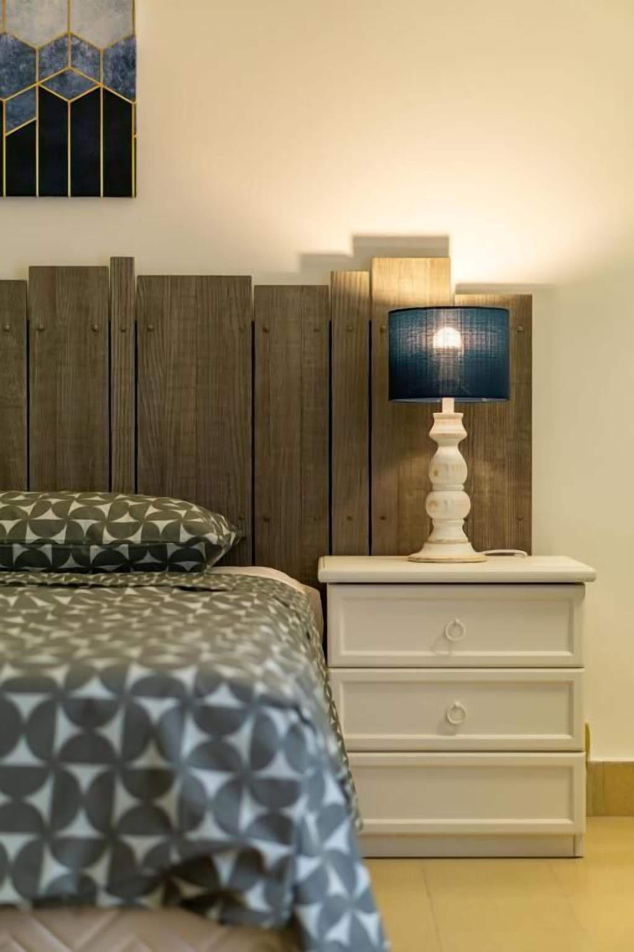 Una cama con una colcha de estampado geométrico, una mesita de noche blanca con cajones y una lámpara fijada a la pared.