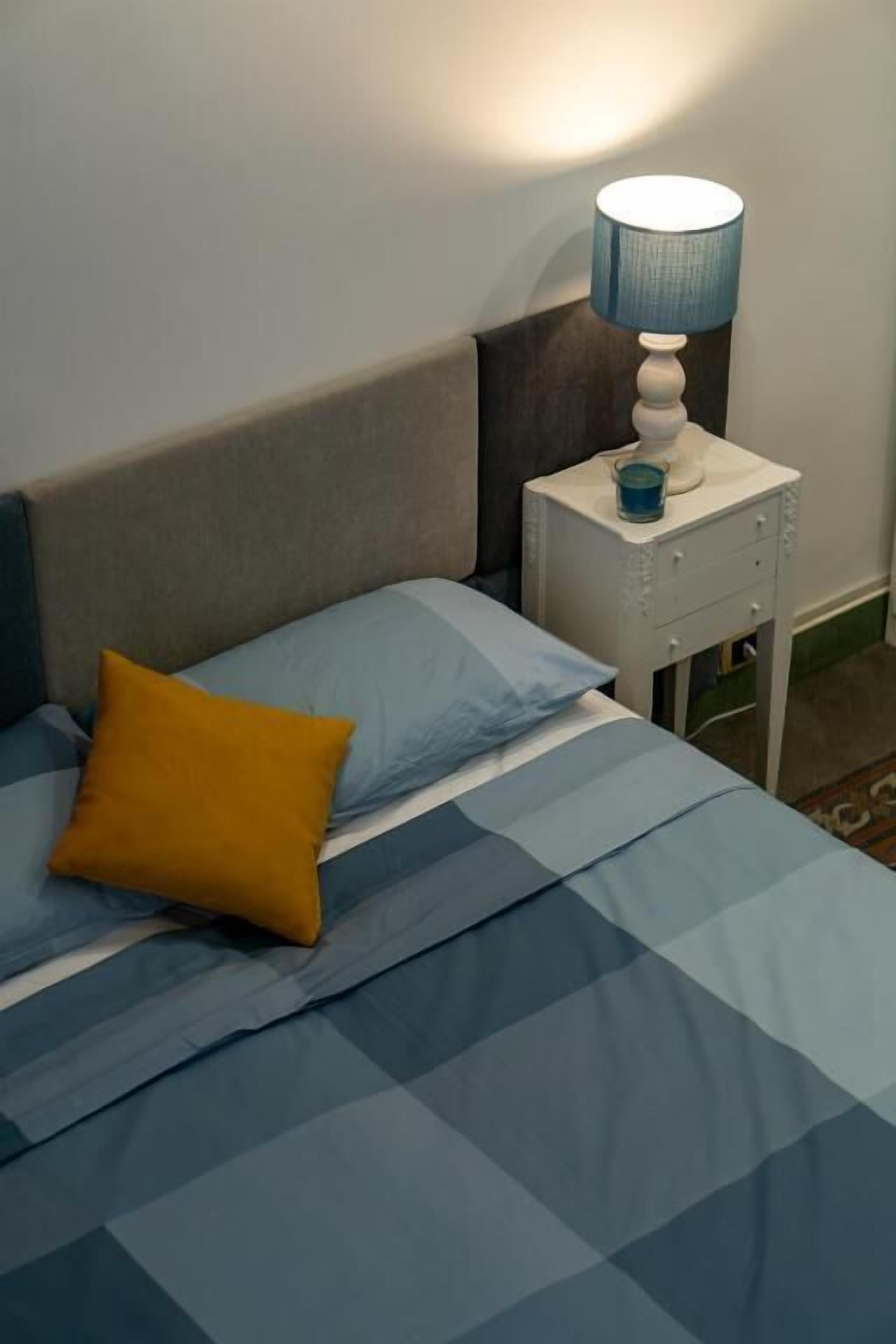 Una cama con ropa de cama azul y un almohadón amarillo, una lámpara de noche y una mesita de noche con una lámpara azul.