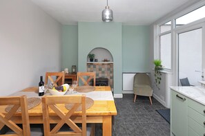 Een eethoek met een tafel gedekt voor twee personen, een wijnfles, glazen en een schaal met fruit. Er is een houten stoel, een beklede bank en een klein plankje met decoratieve objecten.