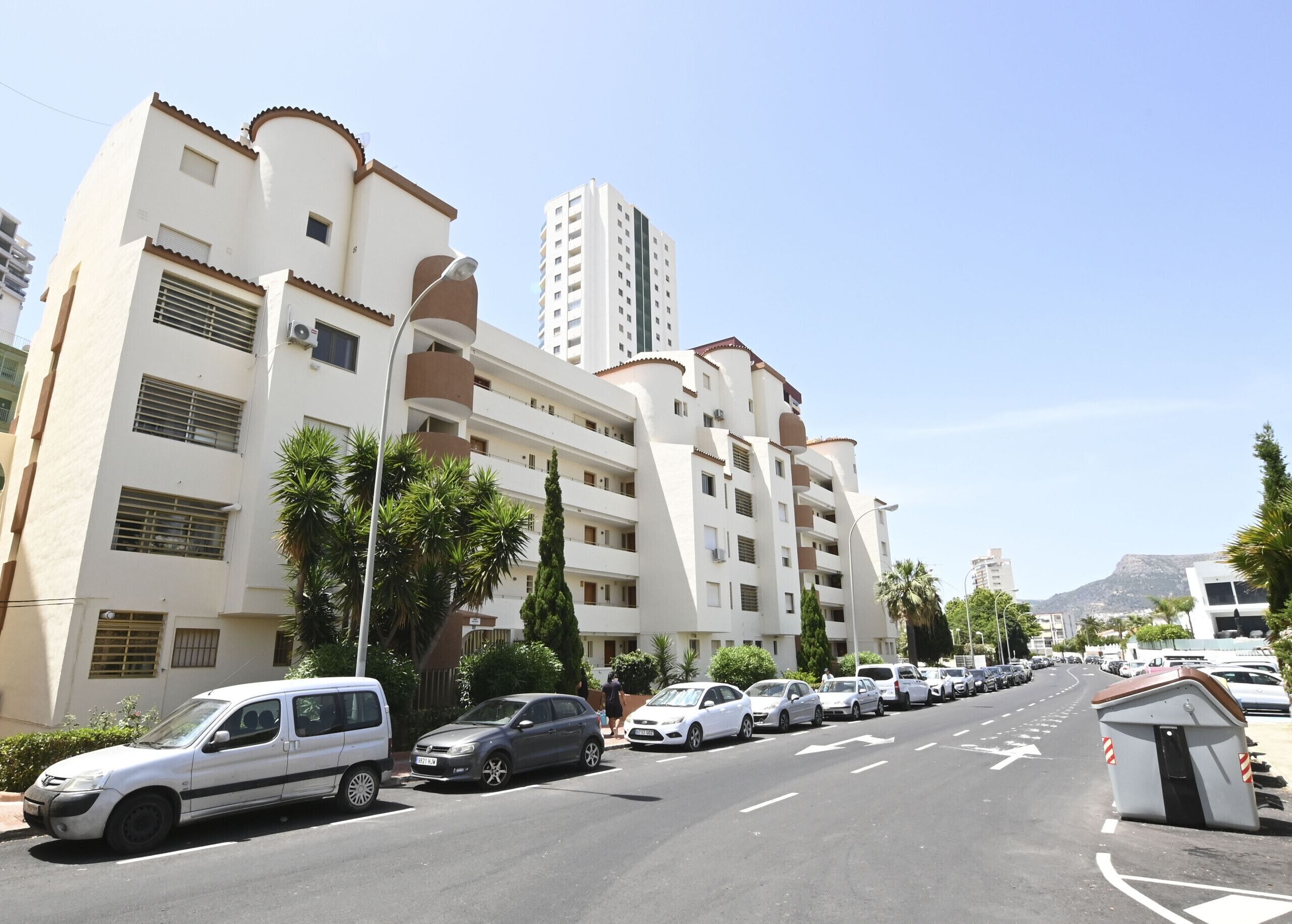 A multi-story residential building with balconies, parked cars, and a clear sky.