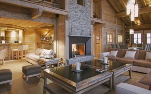 A spacious living area with a stone fireplace, wooden beams, and a modern kitchen in the background. - 6-Bedroom Chalet with Wellness Area (La Clusaz)