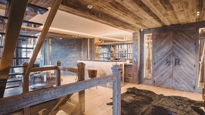 Um interior rústico com vigas de madeira aparente, parede de pedra, área de bar e um tapete de couro grande.
