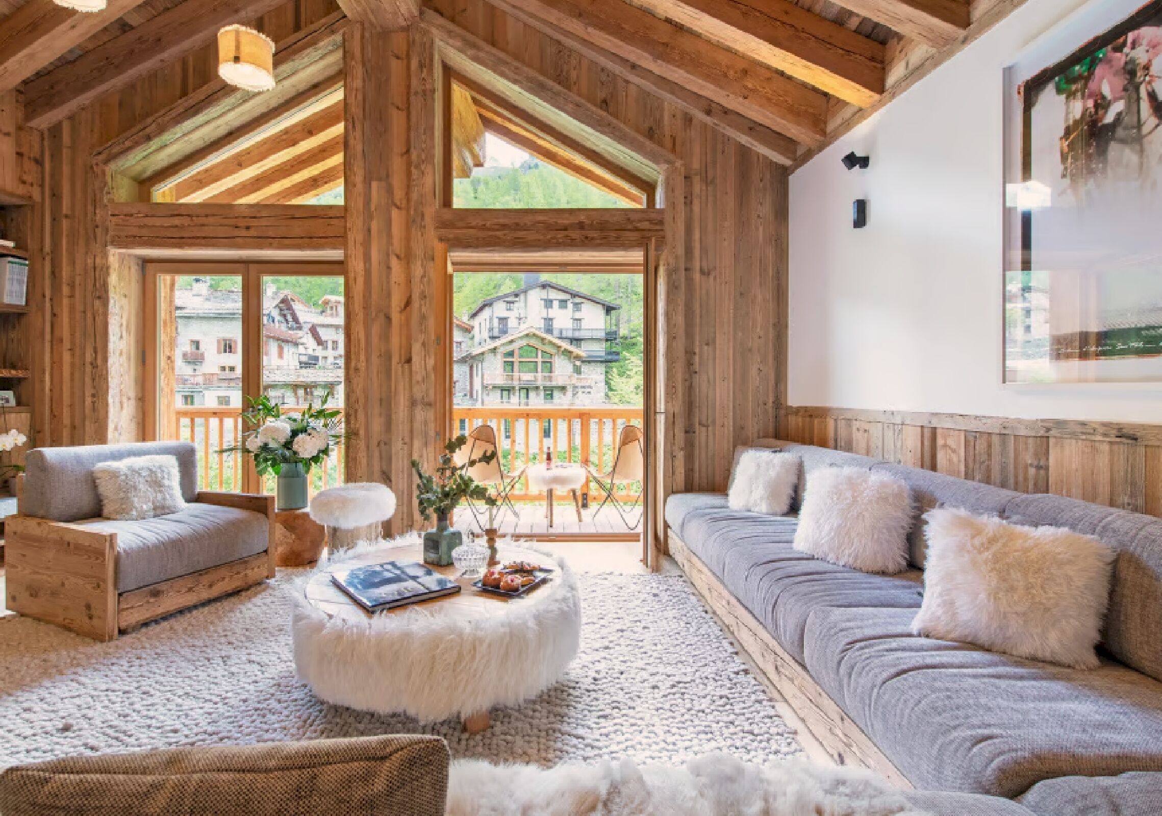 Ein gemütliches Zimmer mit Holzbalkendecke, einer Couch, einem Sessel, einem runden Tisch und Blick auf ein Haus durch große Fenster.