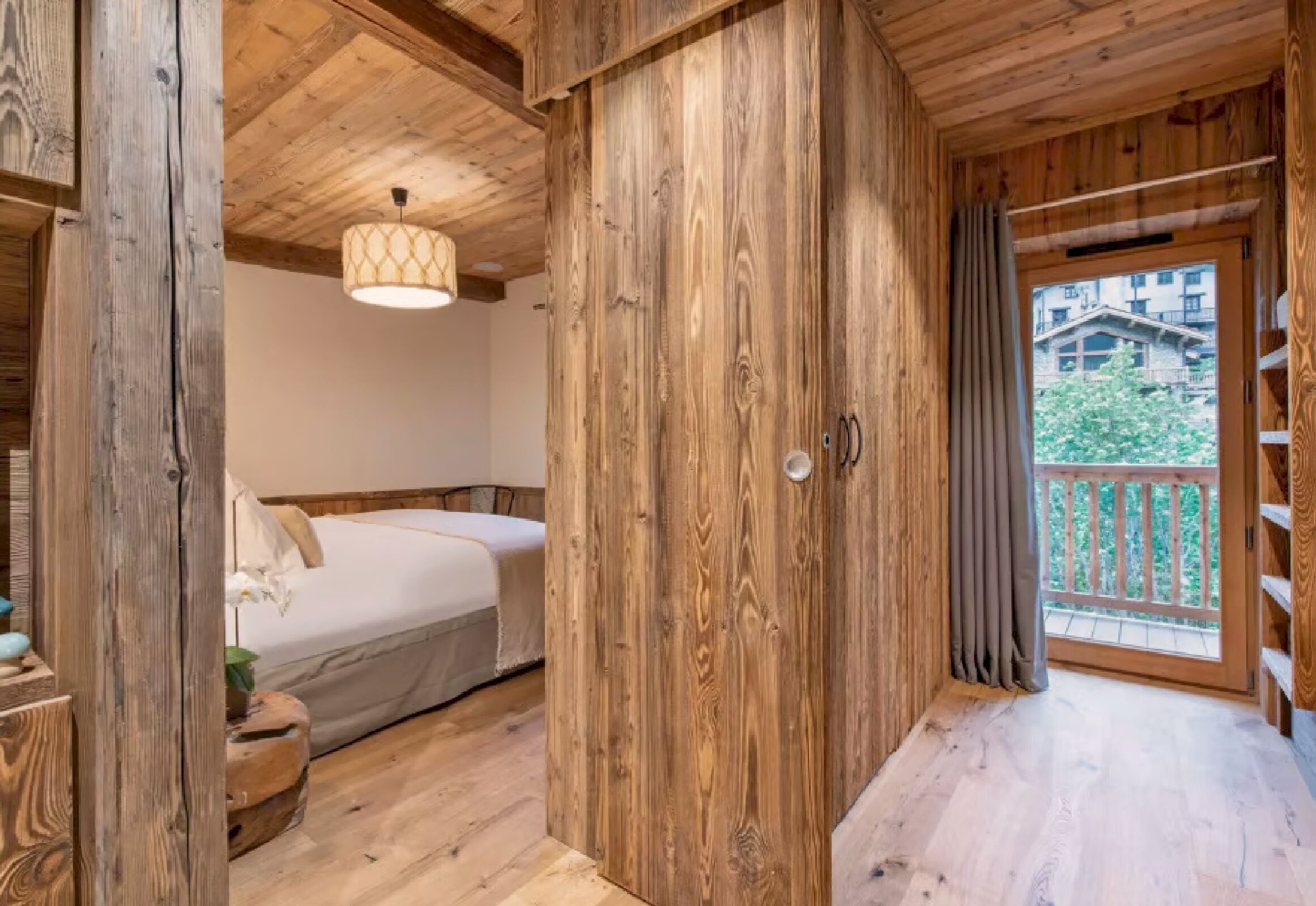A wooden cabin interior with a bed, a hanging light fixture, and a balcony with a view of greenery.