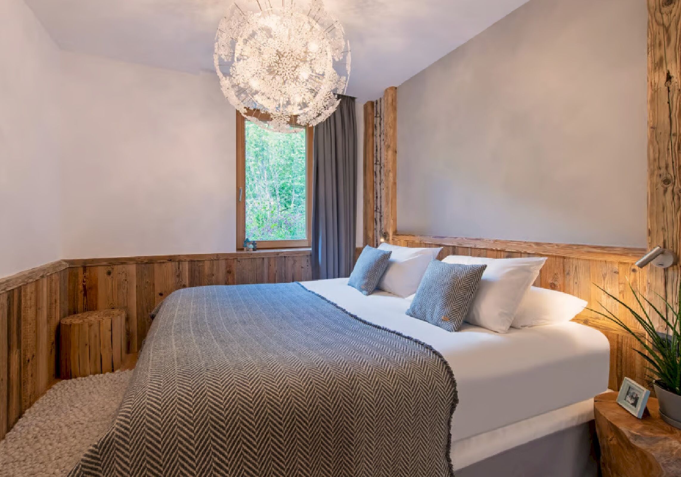 Un dormitorio con una cama grande, cabecera de madera, una araña, una ventana con cortinas y una planta en la mesita de noche.