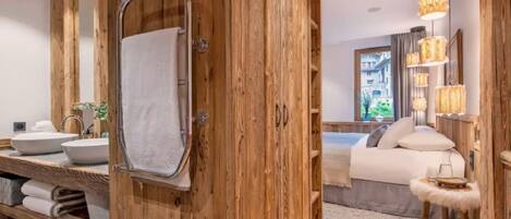 Une porte en bois donne accès à une salle de bain équipée d’un miroir, de deux lavabos et d’un porte-serviettes.