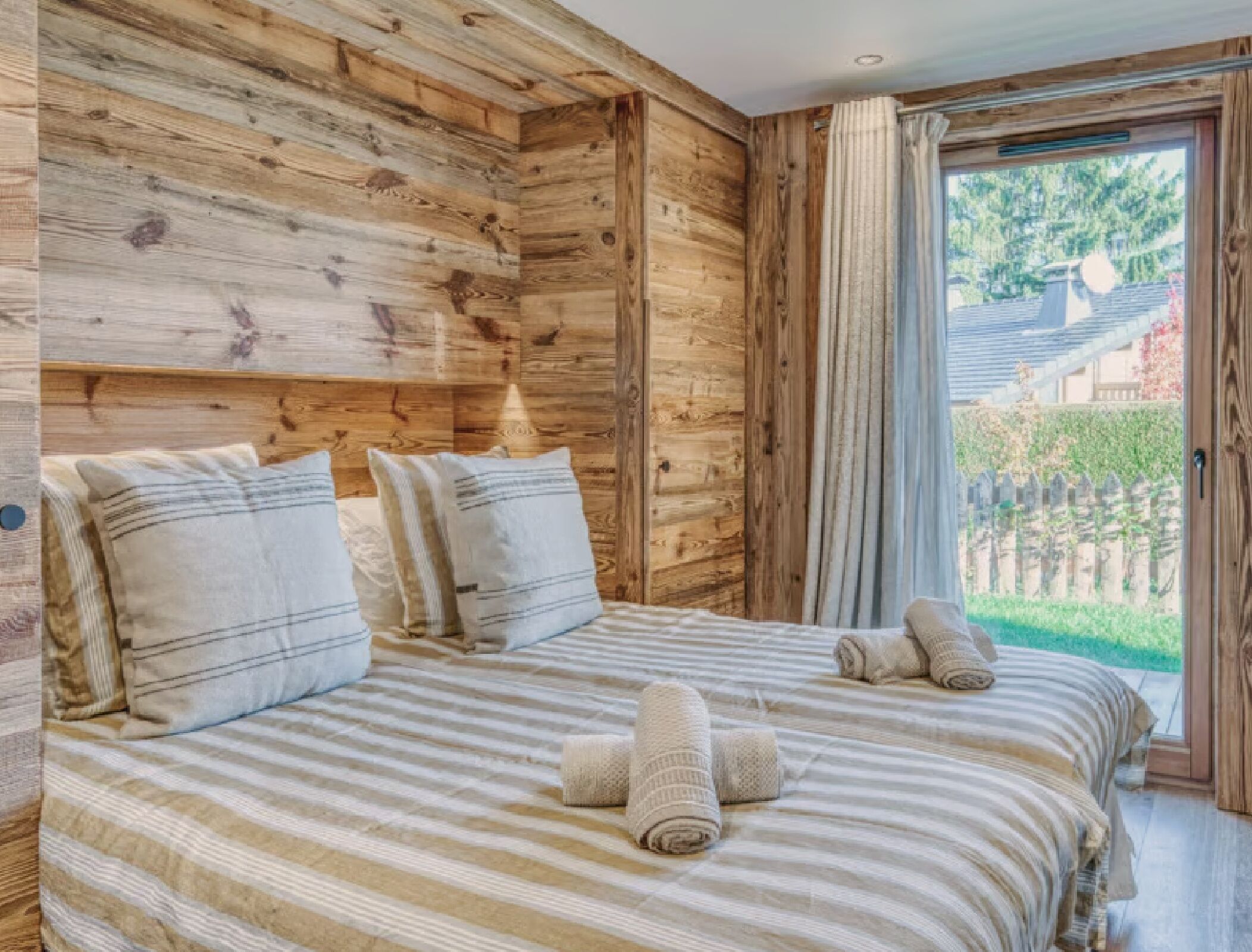 Une chambre à coucher avec une tête de lit en bois, une grande fenêtre avec des rideaux, une couvre-lit à rayures et des serviettes enroulées sur le lit.