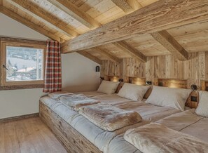 Une pièce avec un plafond en bois, un lit recouvert de plusieurs coussins, une fenêtre donnant sur un paysage enneigé et un rideau à carreaux.