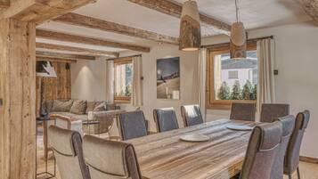 Ein Essbereich mit Holztisch, Ledersesseln, Holzbalken an der Decke und großen Fenstern mit Vorhängen.