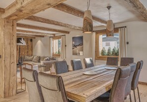 Een eethoek met een houten tafel en lederen stoelen, houten balken in het plafond en grote ramen met gordijnen.