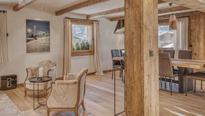 Ein rustikales Interieur mit freiliegenden Holzbalken, einem Essbereich mit Tisch und Stühlen sowie Blick auf eine verschneite Landschaft durch ein Fenster.