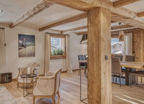 Ein rustikales Interieur mit freiliegenden Holzbalken, einem Essbereich mit Tisch und Stühlen sowie Blick auf eine verschneite Landschaft durch ein Fenster.