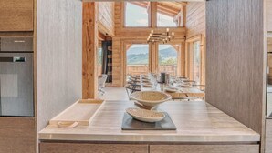 A modern kitchen with wooden cabinetry, a central island, and a view of a dining area with a table set for a meal. - Modern Chalet with Panoramic Views (Les Avanchers-Valmorel)