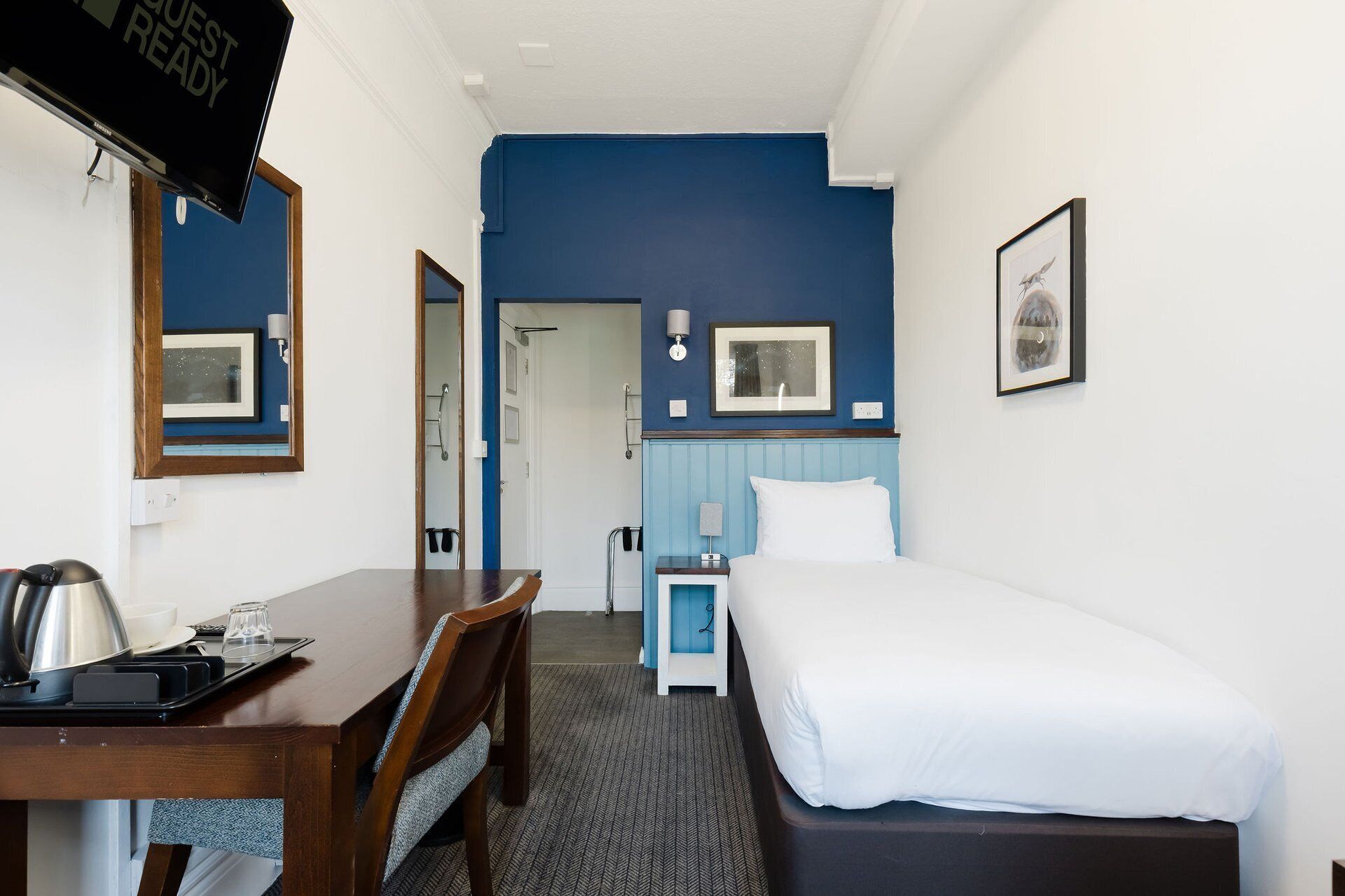 Habitación de hotel con cama, escritorio con tetera, silla, mesita de noche pequeña y televisor mostrando 'GUEST READY'.